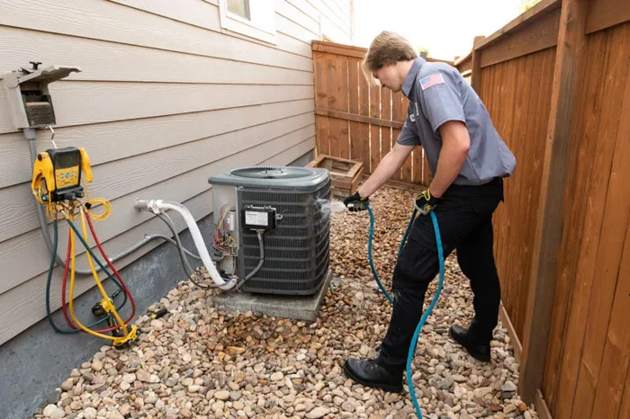 A Done technician rinses an outdoor AC condenser unit with a blue hose against home siding in Aurora, Colorado.