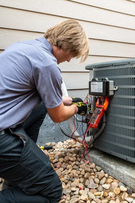 Done technician kneeling at an outdoor condenser testing components with a multimeter during an air conditioner repair in Denver, Colorado.