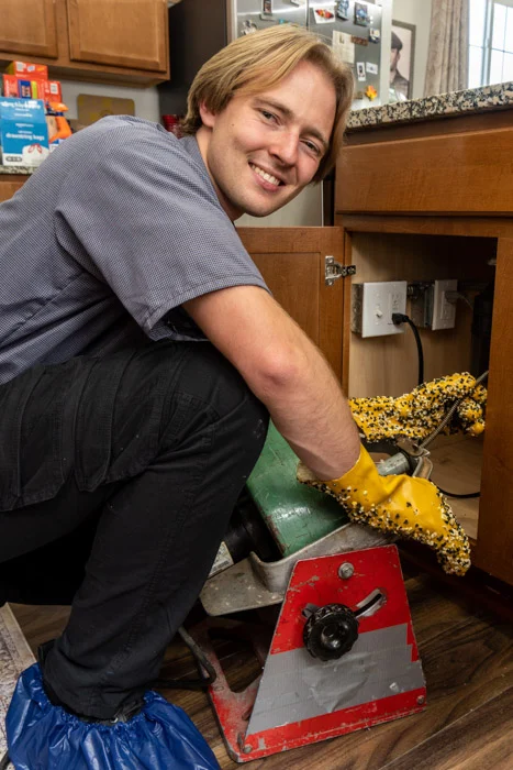 Done plumber in gloves and shoe covers smiling while running a drain cleaning machine beneath a kitchen sink in an Aurora home.