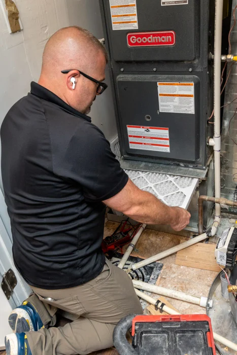 Done HVAC technician kneeling to change the air filter on a Goodman furnace in a Denver home utility closet.
