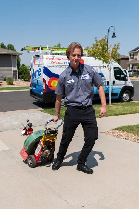 A Done technician stands holding a drain cleaning machine in front of a Done service van on a driveway in Denver, Colorado.