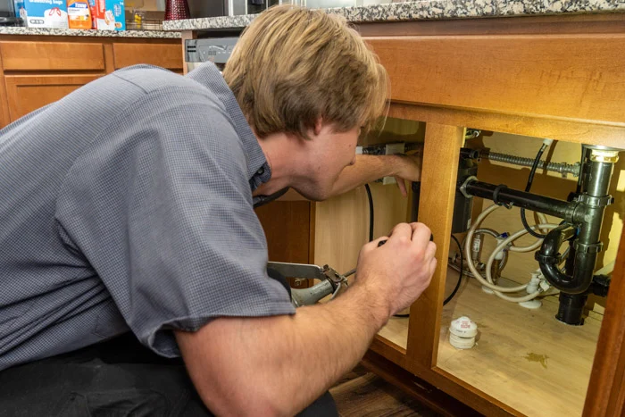Done plumber using pliers to work on the drain piping under a kitchen sink cabinet in an Aurora home.