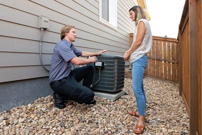 Done technician kneeling beside an outdoor condenser unit talking with a homeowner along the side of a house in Denver, Colorado.