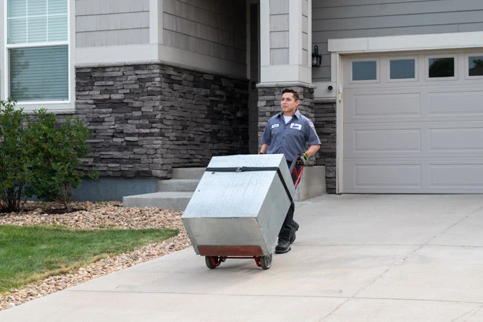 A Done technician moves a metal furnace unit on a hand truck up a driveway past a garage at a Denver, Colorado home.