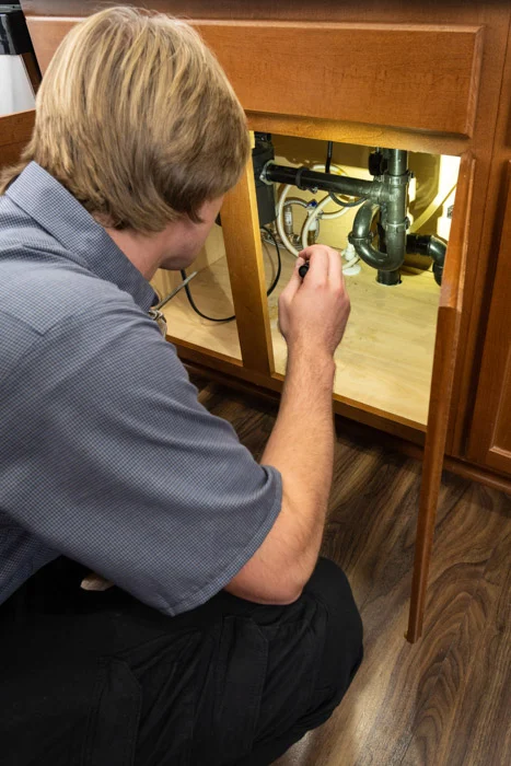 Done plumber crouched at an open cabinet adjusting the drain and supply lines beneath a kitchen sink in a Denver home.