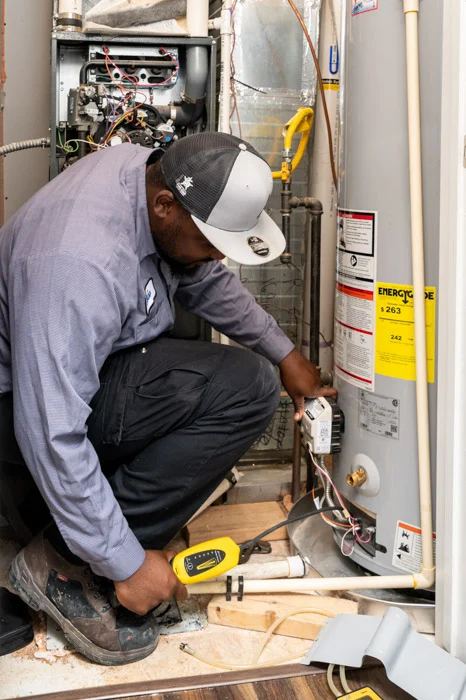 Done technician kneeling to service the gas control valve on a water heater beside an open furnace in an Aurora home.