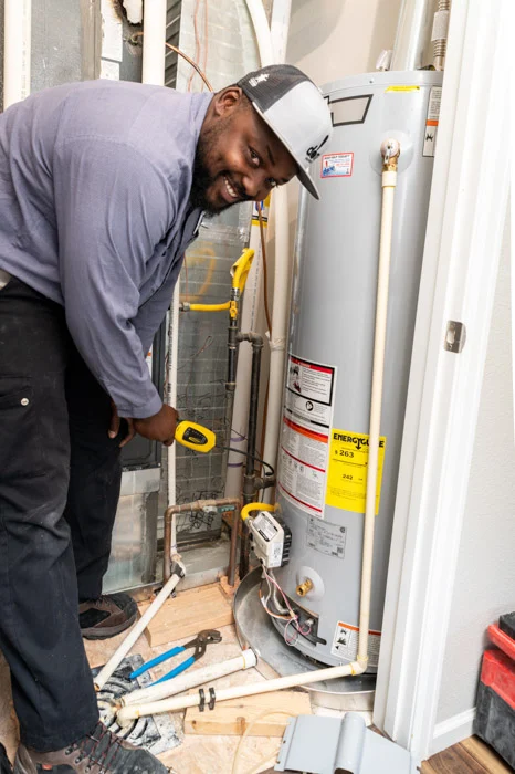 Smiling Done technician with a tester inspecting a gas water heater beside HVAC equipment in an Aurora home.