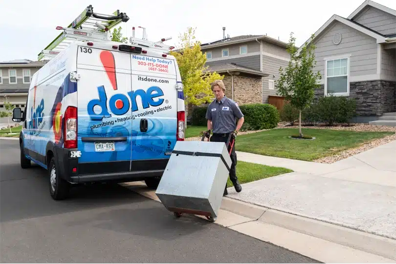 A Done HVAC technician pushing a furnace on a dolly past their work vehicle.