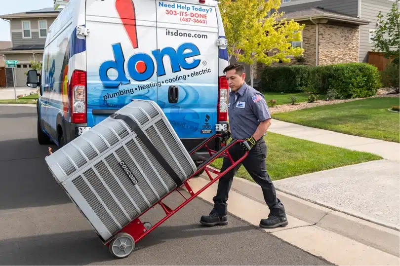 A Done HVAC technician pushes a dolly with an AC unit from their truck to a home