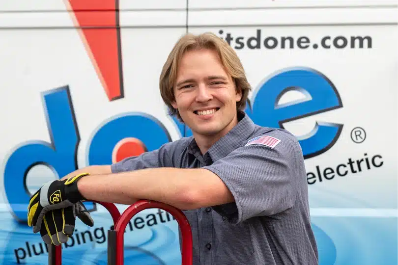 A Done technician smiles at the camera while leaning against a dolly carrying an HVAC system