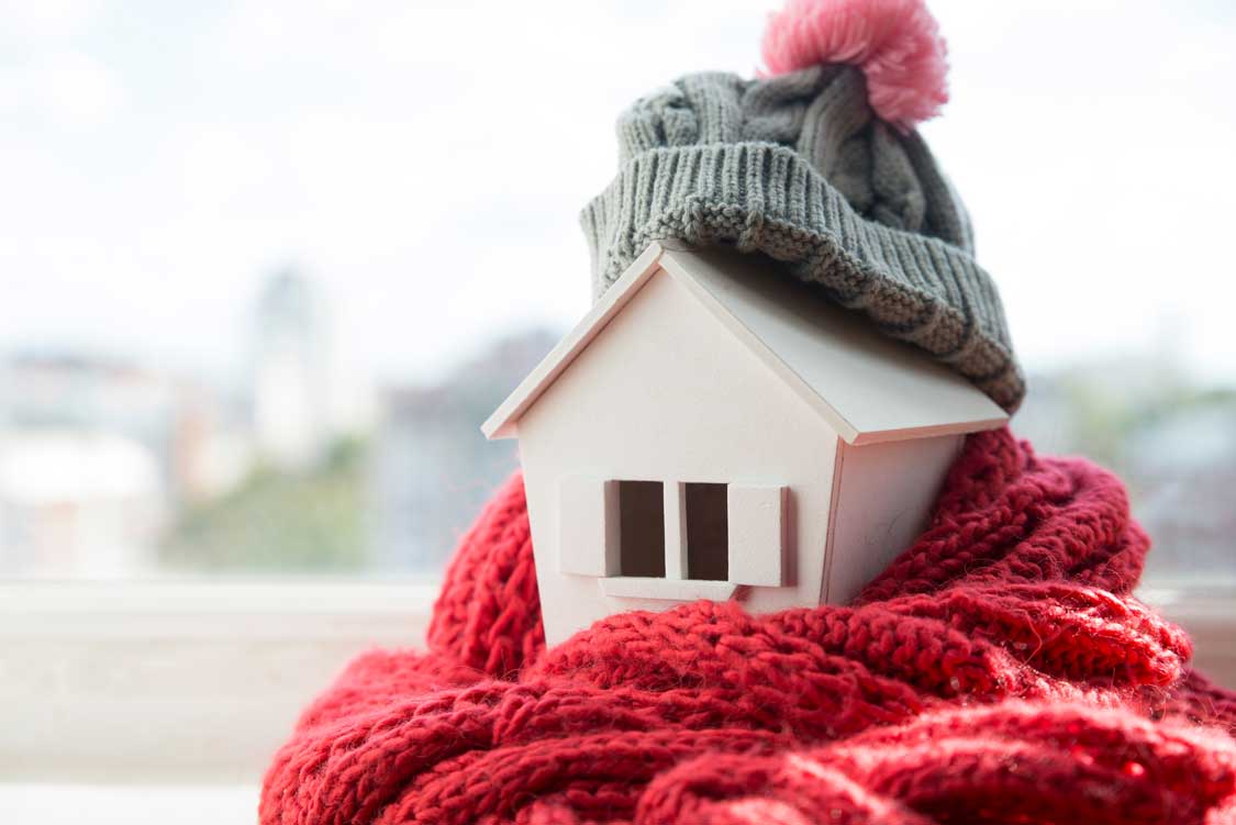house in winter - heating system concept and cold snowy weather with model of a house wearing a knitted cap